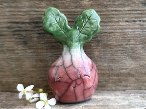 a very smiley and happy radish spirit raku ceramic sculpture. it has a round body glazed in a soft radish red colour. the top of its body fades to white, and it has two green leaves on the top of its head. body. it is round with green leaves on top!