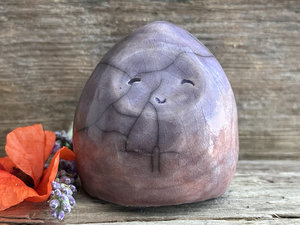 a small, kind of dome or egg-shaped raku ceramic sculpture of a woodland nature spirit. it has a happy, gentle face, and is glazed in a top-to-bottom gradient of muted violet to a soft, warm orangey mauve colour. (it’s actually really hard to describe the colours on this one as they all kind of overlap, but those are the main ones.)