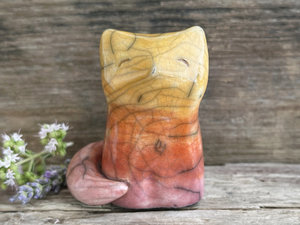Raku ceramic sitting fox glazed in a gradient from mauve to orange to golden yellow (bottom to top). It has a kind face, and its tail is curled around its body.