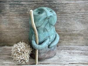 raku ceramic nature spirit warrior sculpture. it is glazed in a gradient from warm brown to forest green (bottom to top) and carries a stick in one of its arms. it has a sad and worried but determined look on its gentle face.