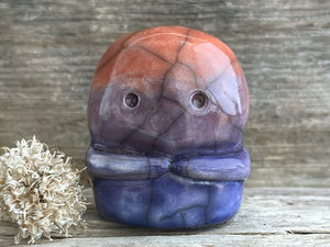 A kodama nature spirit ceramic guardian statue glazed in a bottom-to-top gradient of indigo to violet to mauve to red-orange. It has a wide face, two big eyes, and cute little arms.