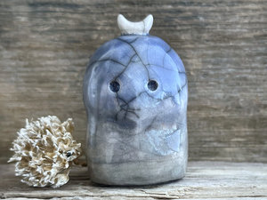A kodama nature and forest spirit kodama kami ceramic raku sculpture glazed in a bottom-to-top gradient of soft grey-brown to blue. It has a gentle face, and a white crescent moon on its head.