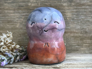 A smiley and loving Jizo Shinto raku ceramic sculpture talisman glazed in a bottom-to-top gradient of red-orange to mauve to purple to blue. It has a kind, gentle face and is a wonderful smooth shape to hold in the hand.