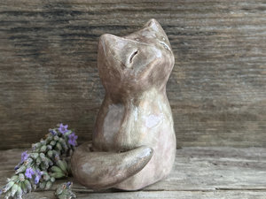 a beautiful Shinto Inari kitsune fox kami spirit ceramic raku statue. it's looking up with a peaceful, happy expression on its gentle face and is glazed in soft browns and pink-browns with its tummy being lighter than the rest. its tail is curled around its body on its right side, and it has white, snow-like marks in the glaze on the left side of its head and back.