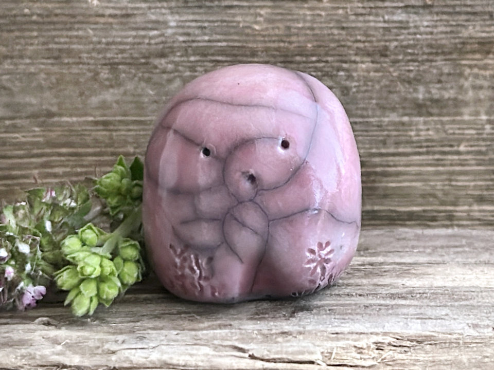 a small and stout nature spirit raku sculpture glazed in mauve pink. it has a cute little face, and little plant carvings on its front.