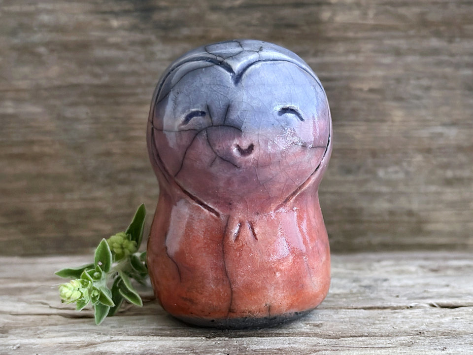 a smiley and loving Jizo Shinto raku ceramic sculpture talisman glazed in a bottom-to-top gradient of soft red-orange to purple to blue. she has a heart shape around her kind, gentle face and is a wonderful smooth shape to hold in the hand.
