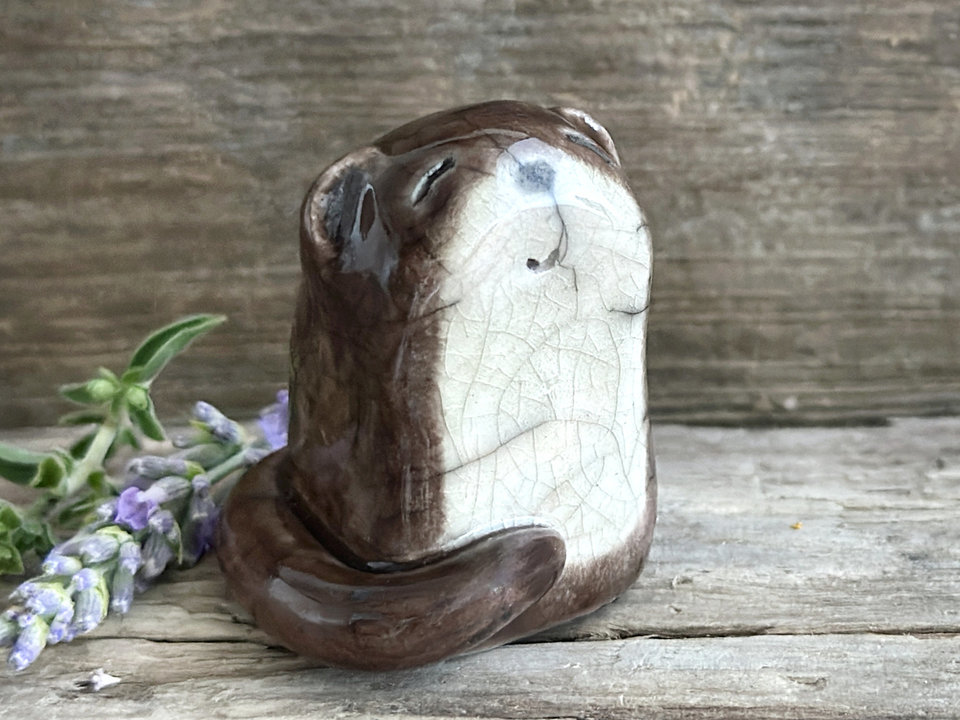 Adorable river otter raku sculpture glazed in brown with a cream-coloured tummy. It has a black nose, little ears, and a tail wrapped around its body. It also has a kind and gentle face.