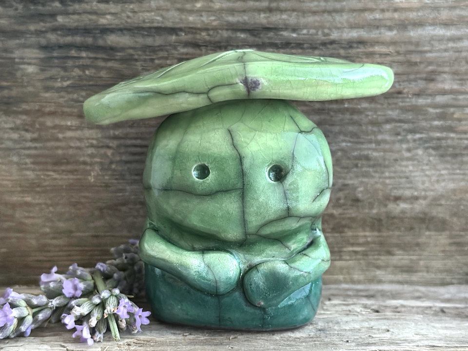 Raku ceramic sculpture glazed in forest green to spring green (bottom to top) with a kind, but slightly intense, face, and a brighter yellow-green leaf on its head. It also has two cute little arms.