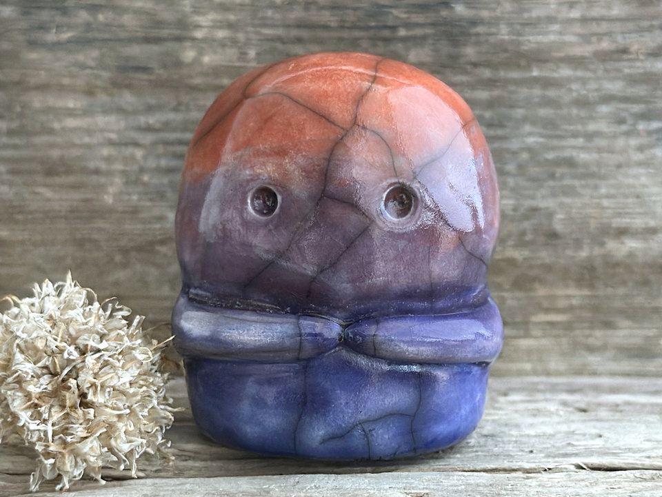 A kodama nature spirit ceramic guardian statue glazed in a bottom-to-top gradient of indigo to violet to mauve to red-orange. It has a wide face, two big eyes, and cute little arms.