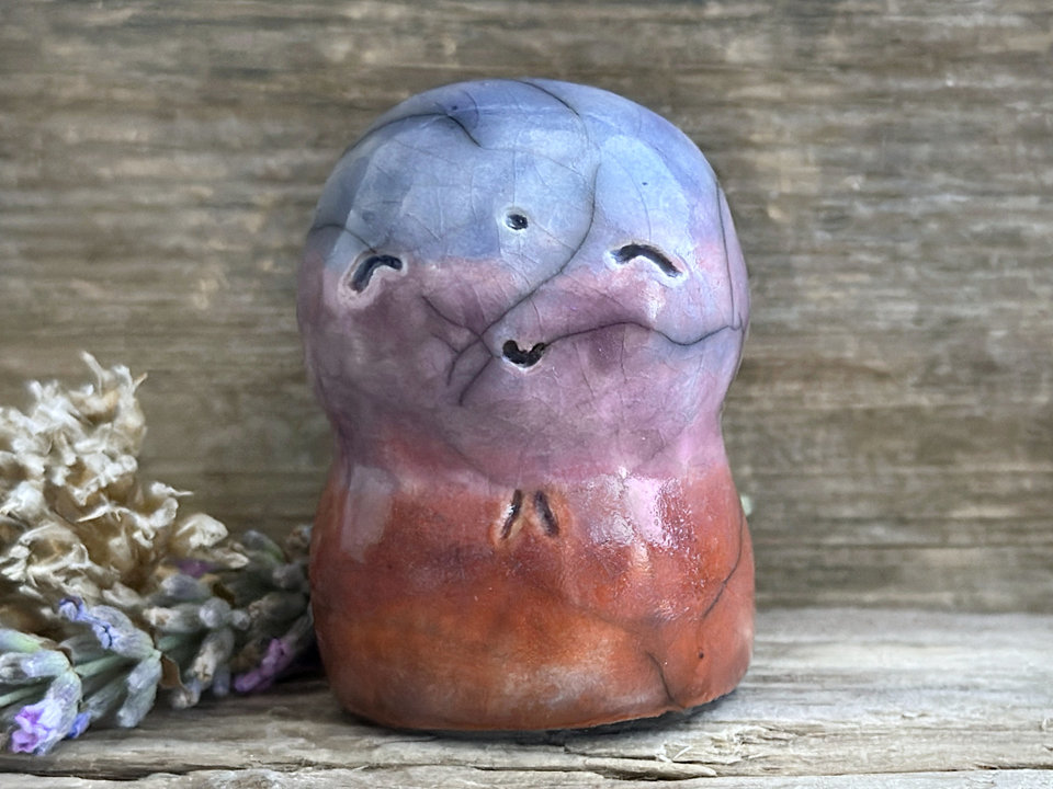 A smiley and loving Jizo Shinto raku ceramic sculpture talisman glazed in a bottom-to-top gradient of red-orange to mauve to purple to blue. It has a kind, gentle face and is a wonderful smooth shape to hold in the hand.