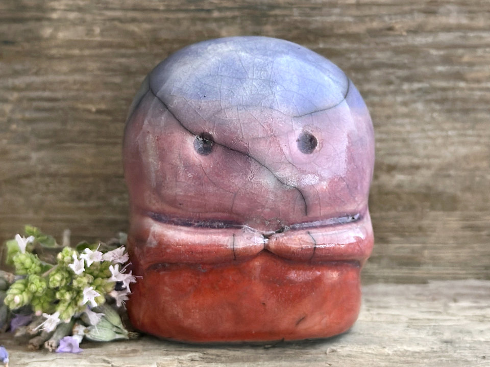 A kodama nature spirit ceramic guardian statue glazed in a top-to-bottom gradient of blue to mauve to red-orange. It has a wide face, two big eyes, and cute little arms.
