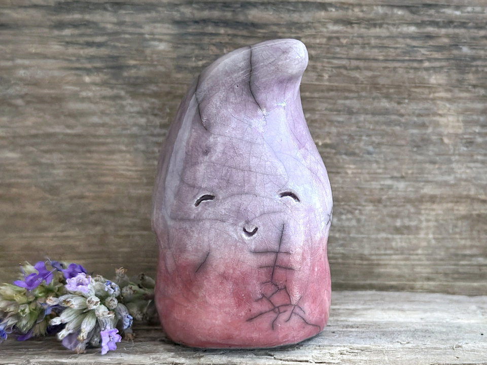 raku ceramic gnome spirit glazed in a bottom-to-top gradient of soft bordeaux red to purple. it has a happy, gentle, kind face, and the top of its head is curled over, a little like it's wearing a nightcap.