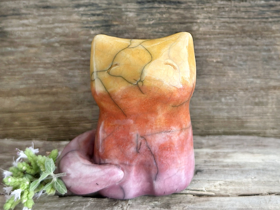 Raku ceramic sitting fox glazed in a gradient from mauve to orange to golden yellow (bottom to top). It has a kind face, and its tail is curled around its body.