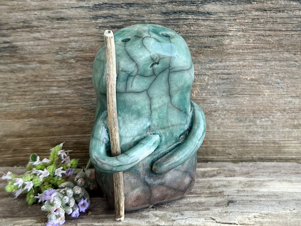 raku ceramic nature spirit warrior sculpture. it is glazed in a gradient from warm brown to forest green (bottom to top) and carries a stick in one of its arms. it has a sad and worried but determined look on its gentle face.