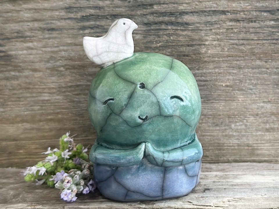 a shinto jizo nature spirit guardian raku ceramic sculpture glazed in a bottom-to-top gradient of blue to forest green to spring green. they have a happy, smiley face, little praying hands, and a sweet white bird on their head.