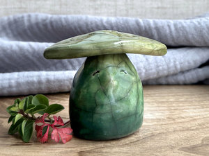 Raku ceramic sculpture glazed in a bottom-to-top gradient of forest green to green to spring green with a kind, contented face. It has a yellow-green leaf on its head.