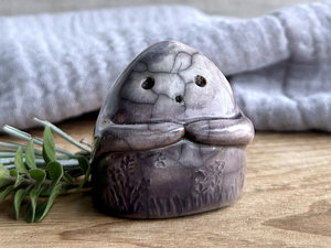 vaguely dome-shaped nature spirit ceramic sculpture. it is glazed dark violet and lighter grey-violet colours and has plant and flower carvings on its body. it also has a gentle face, and two little arms across its front.
