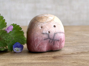 a small and stout nature spirit raku sculpture glazed in peach. it has a cute little face, and little plant carvings on its front. there are tiny dark grey spiderweb patterns in the glaze, in addition to the normal raku crackles, from where raindrops fell on it.