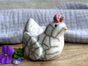 a white raku ceramic chicken with grey eyes, a pale orange beak, and a bordeaux red comb on the top of her head. she looks very sweet.