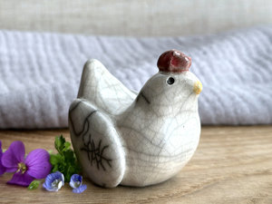 a white raku ceramic chicken with grey eyes, a pale orange beak, and a bordeaux red comb on the top of her head. she looks very sweet.