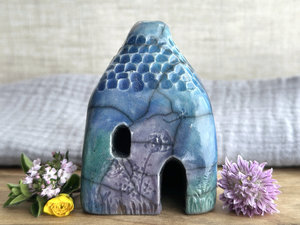 Raku ceramic kurinuki cottage glazed in forest green, blue, indigo, and violet. It has a cute little chimney, plant carvings on its walls, a cute door and lots of little windows, one of which is in the shape of a crescent moon.