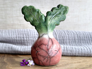 a very smiley and happy radish spirit raku ceramic sculpture. it has a round body glazed in a soft radish red colour. the top of its body fades to white, and it has two green leaves on the top of its head/body. there are some small, round, marks/depressions on its face on its right side.