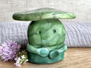 Raku ceramic sculpture glazed in forest green to spring green (bottom to top) with a kind, but slightly intense, face, and a brighter yellow-green leaf on its head. It also has two cute little arms.