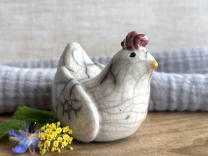 a white raku ceramic chicken with grey eyes, a pale orange beak, and a bordeaux red comb on the top of her head. she looks very sweet.