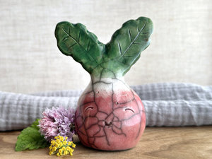 a very smiley and happy radish spirit raku ceramic sculpture. it has a round body glazed in a soft radish red colour. the top of its body fades to white, and it has two green leaves on the top of its head/body. there are some tiny glaze disturbance marks on the top of its head that make it look like it's been in the ground.