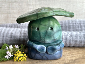 Raku ceramic sculpture glazed in a bottom-to-top gradient of blue to forest green to spring green with a kind, but slightly intense, face, and a spring green leaf on its head. It also has two cute little arms.