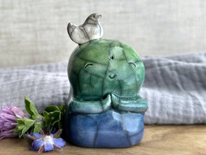 a raku ceramic jizo nature spirit guardian sculpture glazed in a bottom-to-top gradient of blue to forest green to spring green. they have a happy, smiley face, little praying hands, and a sweet white bird on their head.