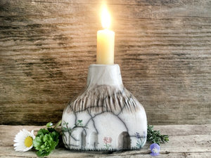 Raku ceramic candlestick in the shape of a rectanglar cottage. The top is brown and carved to look like thatch. The rest is a white cottage with windows, a door, and flower and plant carvings.