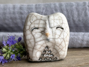 Raku ceramic little owl or owlet kami nature spirit statue with sort of pointed ears. It’s glazed in white and grey-brown with gentle, smiling eyes and a little golden yellow beak.