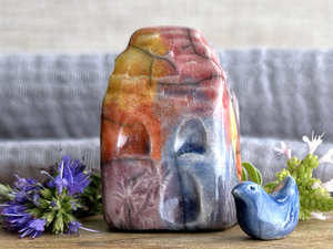 Tiny raku ceramic cottage glazed in golden yellow, warm red-orange, bordeaux red, blue, and purple. It's carved to look like slates. It has a carved door and windows and lots of plant and flower carvings too, and it comes with the little blue bird that's sitting next to it.