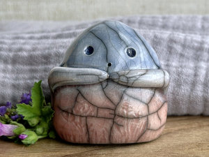 a vaguely dome-shaped nature spirit ceramic sculpture. it is glazed in a bottom-to-top gradient of soft, pale reddish pink to pale blue and has plant and flower carvings on its body. it also has a gentle face, and two little arms across its front.