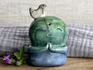 a raku ceramic jizo nature spirit guardian sculpture glazed in a bottom-to-top gradient of blue to forest green to spring green. they have a happy, smiley face, little praying hands, and a sweet white bird on their head.
