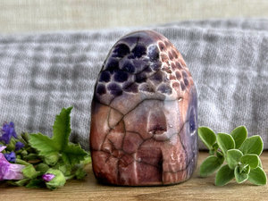 Tiny raku ceramic cottage glazed in warm red-orange, bordeaux red, mauve, and purple. Its roof is carved to look like slates. It has a carved door and windows and lots of plant and flower carvings on its walls.