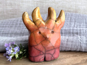 a raku ceramic creature. it has four stubby legs and gets wider as its body goes up. it eventually ends in four small antlers. on the way, it has a gentle face with a cute little nose. it's glazed in a bottom-to-top gradient of red-pink to warm orange to golden yellow for the antlers. it is a friend.