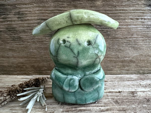 Raku ceramic sculpture glazed in forest green to spring green (bottom to top) with a sweet face and a lighter, yellow-green leaf on its head. It also has two cute little arms.