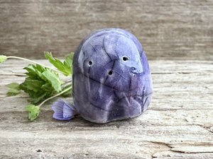 a small and stout nature spirit raku sculpture glazed in violet. it has a cute little face, and a little plant carving on its left side.