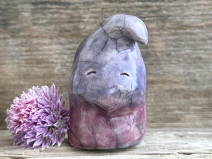 raku ceramic gnome spirit glazed in a bottom-to-top gradient of purple-mauve to indigo-violet. it has a gentle, sleepy, kind face, and the top of its head is curled over, a little like it's wearing a nightcap.