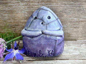 a vaguely dome-shaped nature spirit ceramic sculpture. it is glazed in a bottom-to-top gradient of violet to blue and has plant and flower carvings on its body. it also has a gentle face, and two little arms across its front.