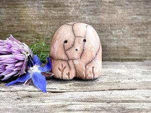 a small and stout nature spirit raku sculpture glazed in peach. it has a cute little face, and a little plant carving on its right side.