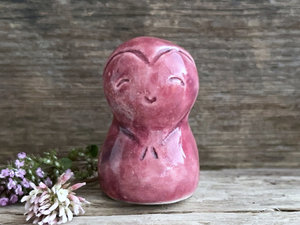 A smiley and loving Jizo Shinto raku ceramic sculpture talisman glazed in warm, pinky red colour with hints of purple. It has a heart shape carved around its kind, gentle face and is a wonderful smooth shape to hold in the hand.
