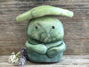 Raku ceramic sculpture glazed in forest green to spring green (bottom to top) with a kind, but slightly intense, face, and a brighter yellow-green leaf on its head. It also has two cute little arms.