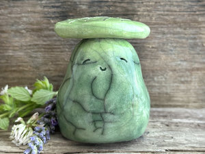 Raku ceramic sculpture who looks a bit like a pear glazed in a mix of forest and spring greens with a gentle, happy face, and a brighter yellow-green leaf on its head.