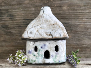 Rustic raku ceramic kurinuki cottage glazed in white with pale green plant and pale purple flower patterns on the walls. The roof has a chimney and is glazed in pale brown and white to look like thatch. It has a cute little doorway and lots of windows.