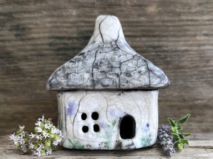 Rustic raku ceramic kurinuki cottage glazed in white with pale green plant and pale violet flower patterns on the walls. The roof has a chimney and is glazed in grey with hints of white to look like slate. It has a cute little doorway and lots of windows.