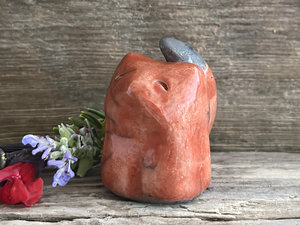 Raku ceramic sitting kitsune fox spirit sculpture glazed in a deep red-orange colour. It has a sweet, gentle face, and its tail points up its back.