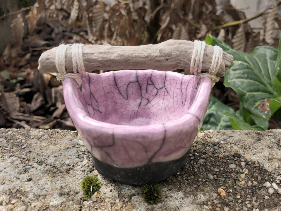 Small raku ceramic bucket with a driftwood handle. The bucket is glazed a mauve pink on the inside and halfway down the outside, and the rest of the outside is left natural, unglazed black.